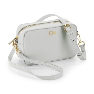 May include: A light gray crossbody bag with gold-colored hardware. The bag has a rectangular shape with a zipper closure and a long, adjustable strap. The initials "EN" are printed in gold on the front of the bag. The bag is made of a smooth material.