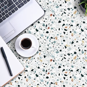 May include: A white laptop computer, a notebook with a black pen, and a white cup of coffee on a terrazzo countertop. The countertop has a white background with black, brown, and blue flecks.