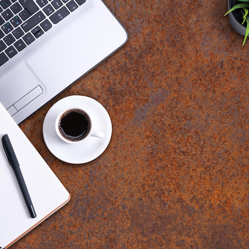 K&ouml;nnte beinhalten: Ein silberner Laptop, ein wei&szlig;es Notizbuch mit einem schwarzen Stift und eine wei&szlig;e Tasse mit schwarzem Kaffee auf einer Untertasse sind auf einer strukturierten, dunkelbraunen, rostigen Metalloberfl&auml;che angeordnet. Eine kleine gr&uuml;ne Pflanze.