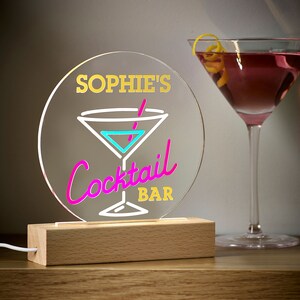 May include: A round, clear acrylic sign with a neon pink and blue cocktail glass design. The sign reads "Sophie's Cocktail Bar" in yellow and white lettering. The sign is lit from behind and sits on a wooden base.