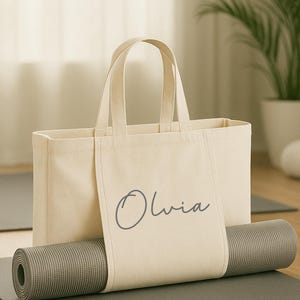 May include: A beige canvas tote bag with the name "Olivia" printed on the front, holding a rolled-up gray yoga mat. The bag has two handles and is sitting on a gray yoga mat.