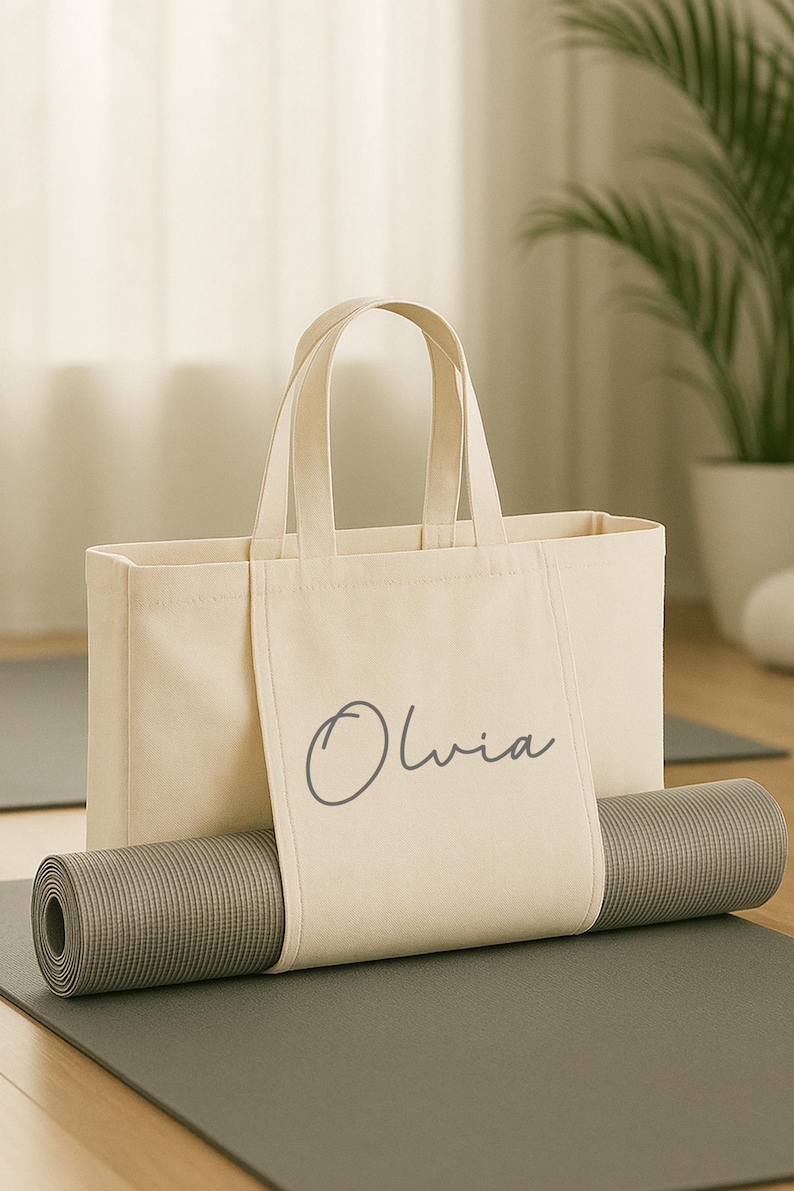May include: A beige canvas tote bag with the name "Olivia" printed on the front, holding a rolled-up gray yoga mat. The bag has two handles and is sitting on a gray yoga mat.