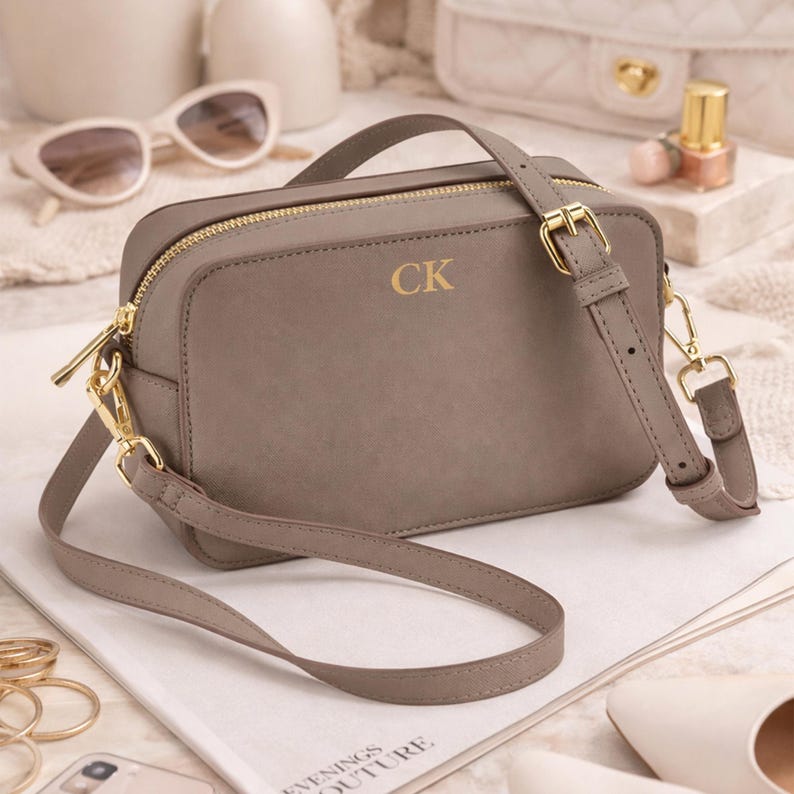 May include: A taupe-colored crossbody bag with gold-tone hardware, including a zipper and buckle. The bag features the gold initials "CK" on the front. It has a long, adjustable strap and a top handle. The bag is sitting on a magazine.