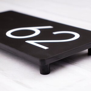 May include: Black house number plaque with white numbers "20" on a black background. The plaque has four small black feet on the bottom.
