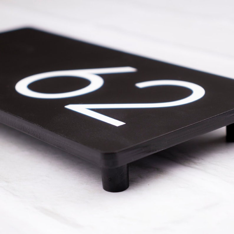 May include: Black house number plaque with white numbers "20" on a black background. The plaque has four small black feet on the bottom.