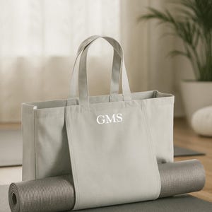 May include: A light gray canvas tote bag with the initials "GMS" in white, holding a rolled-up yoga mat. The mat is gray and has a textured surface. The bag has two handles and a pocket.