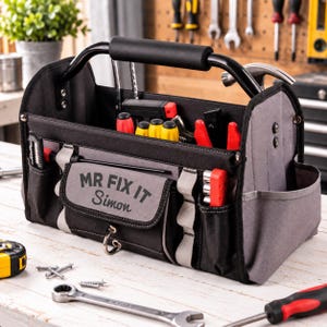 May include: A black and grey open-top tool bag with a metal handle and a front pocket featuring the text MR FIX IT Simon. The bag contains various hand tools including screwdrivers and pliers, with additional tools scattered on the surface.
