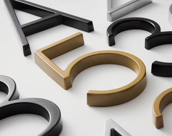 May include: A collection of three-dimensional house numbers and letters in black, gold, and silver finishes. The set includes the letter A, the number 2, the number 3, and the number 5, all featuring a clean, bold, and sans-serif typeface design.