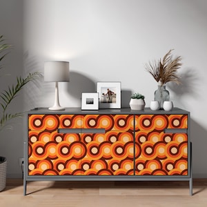 May include: A gray sideboard featuring three cabinet doors with a repeating retro pattern of interlocking circles in shades of orange, brown, and cream. The sideboard has a flat top surface and stands on four thin, straight legs.