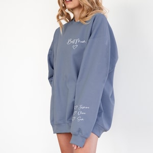 May include: A blue-grey sweatshirt with the text "Best Mum" and a heart graphic on the chest. The sleeve features the names "Jasmine", "Olivia", and "Sam" with heart graphics. The sweatshirt is a relaxed fit.