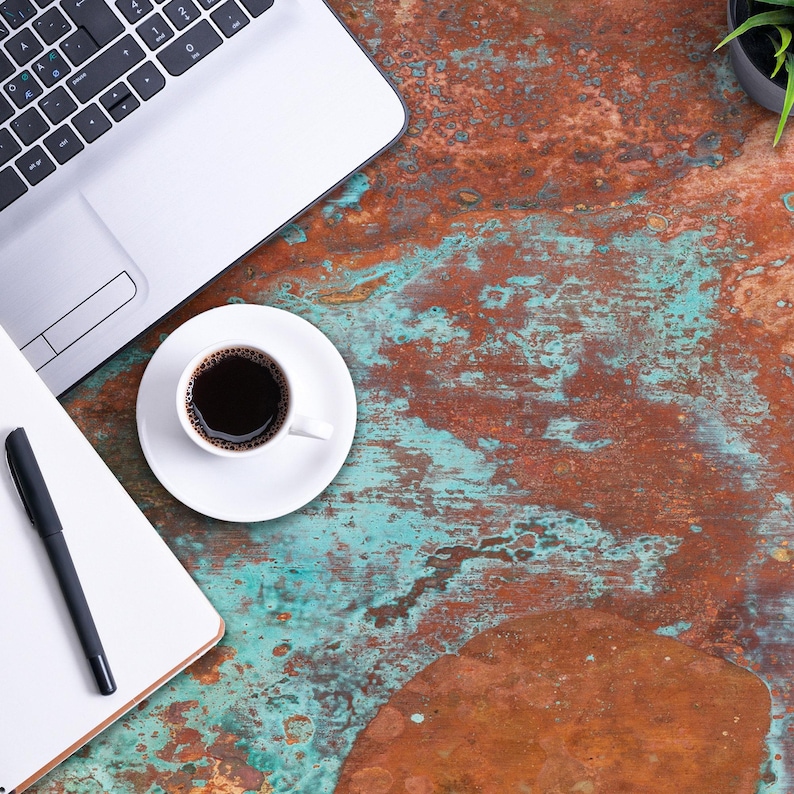 K&ouml;nnte beinhalten: Ein silberner Laptop, ein wei&szlig;es Notizbuch mit einem schwarzen Stift und eine wei&szlig;e Tasse mit schwarzem Kaffee sind auf einer strukturierten Oberfl&auml;che mit oxidierter Kupferoptik in rostorangen und t&uuml;rkisgr&uuml;nen Farbt&ouml;nen angeordnet.