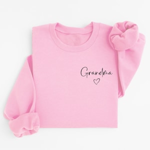 May include: A light pink sweatshirt with the word "Grandma" in black script and a small heart. The sleeves are cuffed, and the sweatshirt is laid flat. The design is simple and elegant, perfect for a gift.