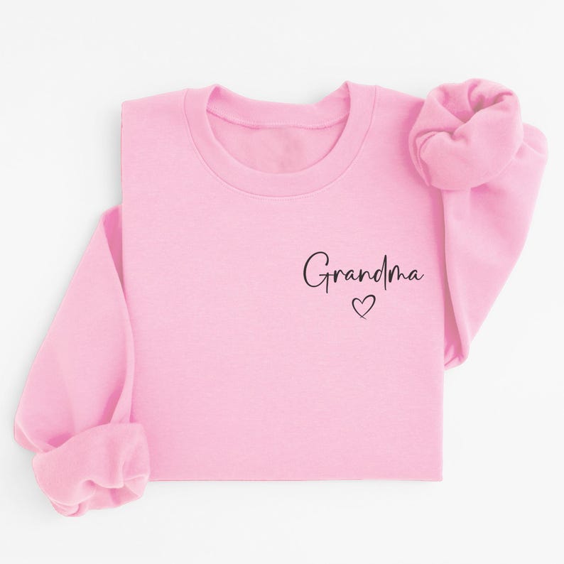 May include: A light pink sweatshirt with the word "Grandma" in black script and a small heart. The sleeves are cuffed, and the sweatshirt is laid flat. The design is simple and elegant, perfect for a gift.