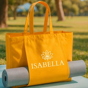 May include: Mustard yellow tote bag with the name "ISABELLA" in white, a lotus flower logo, and a gray yoga mat. The bag has two handles and is sitting on a blue mat, with a park background.