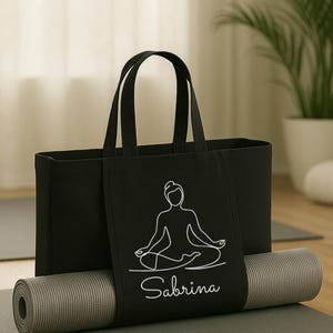 May include: Black yoga tote bag with a white line drawing of a person in a seated yoga pose, with the name "Sabrina" below. A rolled gray yoga mat rests on a gray mat, with a plant in the background.