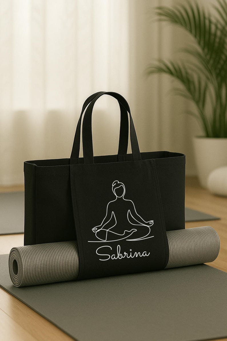 May include: Black yoga tote bag with a white line drawing of a person in a seated yoga pose, with the name "Sabrina" below. A rolled gray yoga mat rests on a gray mat, with a plant in the background.