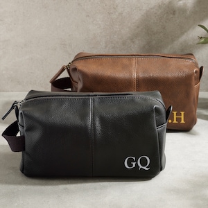 May include: Two leather wash bags with zip fastenings and handles. The bag on the left is black with the initials "GQ" embroidered in white. The bag on the right is brown with the initials "J.H." embroidered in gold.