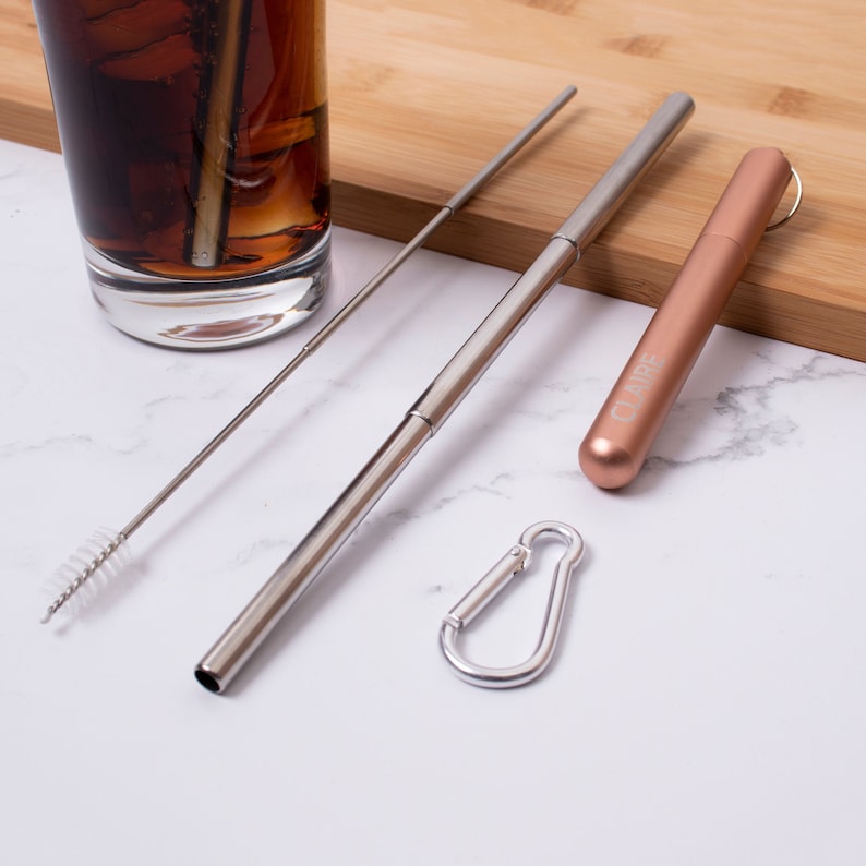 May include: Two silver metal reusable straws with a cleaning brush, a rose gold metal straw case with a carabiner clip, and the word "CLARE" engraved on the case.