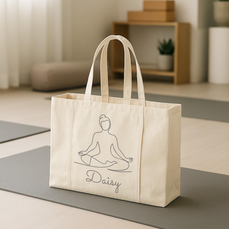 May include: Cream-colored tote bag with a gray line drawing of a person in a yoga pose. The name "Daisy" is written below the drawing in cursive. The bag has two handles and is sitting on a gray yoga mat.