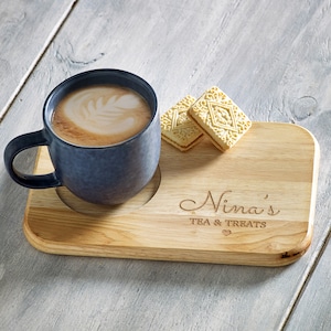 May include: A wooden serving tray with the text "Nina's Tea & Treats" engraved on it. A blue ceramic mug filled with coffee and two white biscuits are on the tray.