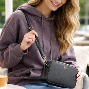May include: Black crossbody bag with gold-tone hardware and a shoulder strap. The bag has a rectangular shape and a zipper closure. The initials "JC" are displayed on the front of the bag. The bag is being held by a person wearing a purple hoodie.