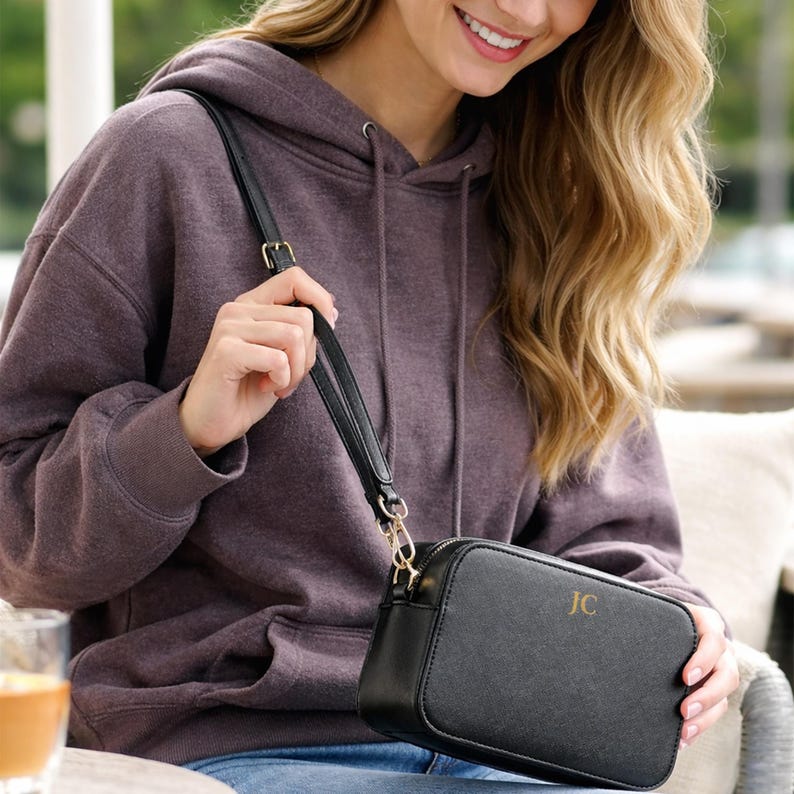 May include: Black crossbody bag with gold-tone hardware and a shoulder strap. The bag has a rectangular shape and a zipper closure. The initials "JC" are displayed on the front of the bag. The bag is being held by a person wearing a purple hoodie.