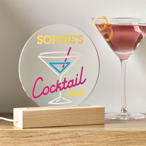 May include: A round, clear acrylic sign with a wooden base. The sign has a pink and blue cocktail glass with a yellow straw and the text "Sophie's Cocktail Bar" in yellow, pink, and white lettering.