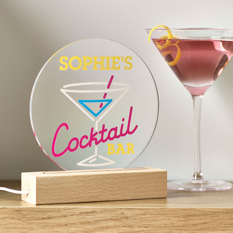 May include: A round, clear acrylic sign with a wooden base. The sign has a pink and blue cocktail glass with a yellow straw and the text "Sophie's Cocktail Bar" in yellow, pink, and white lettering.