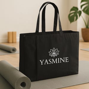 May include: Black tote bag with white text and a lotus flower design. The word "YASMINE" is printed in a clean, sans-serif font. The bag has two handles and is sitting on a gray yoga mat.