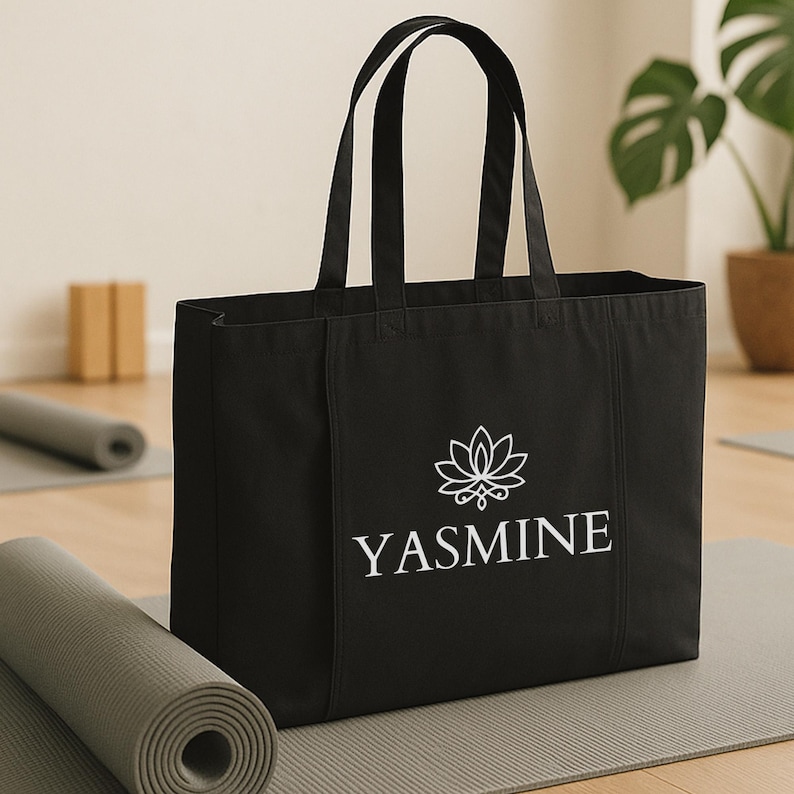 May include: Black tote bag with white text and a lotus flower design. The word "YASMINE" is printed in a clean, sans-serif font. The bag has two handles and is sitting on a gray yoga mat.