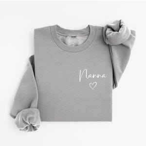 May include: A gray sweatshirt with the word "Nanna" in white script and a small heart. The long-sleeved top is folded, with the sleeves rolled up. The sweatshirt is made of a soft material.