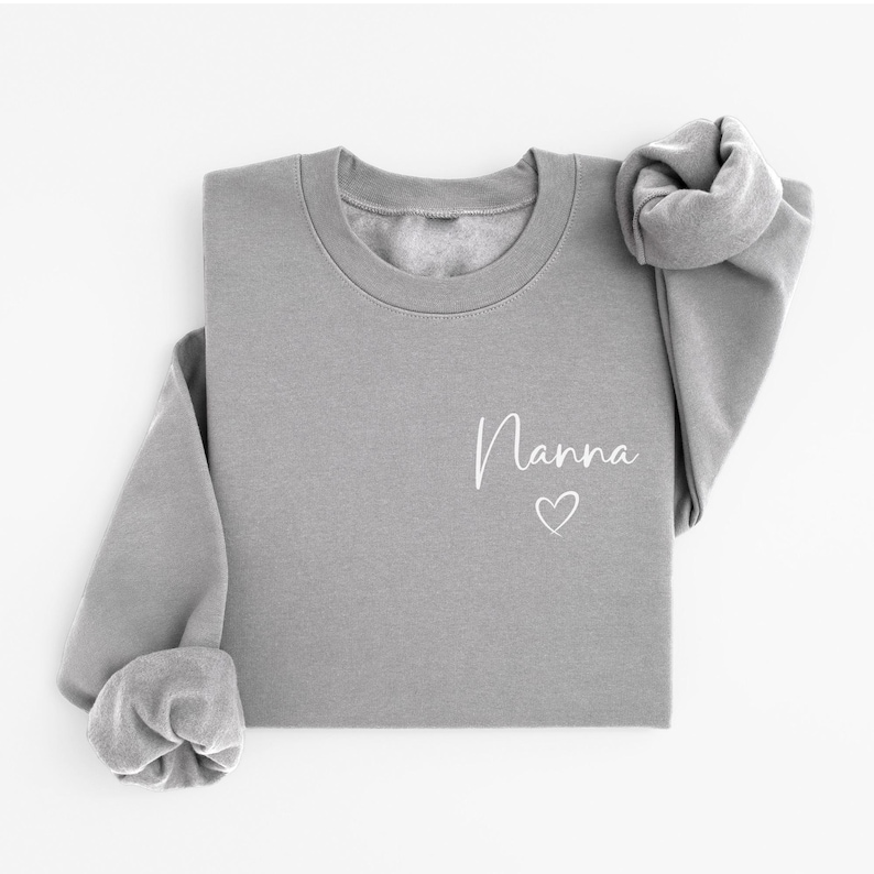 May include: A gray sweatshirt with the word "Nanna" in white script and a small heart. The long-sleeved top is folded, with the sleeves rolled up. The sweatshirt is made of a soft material.