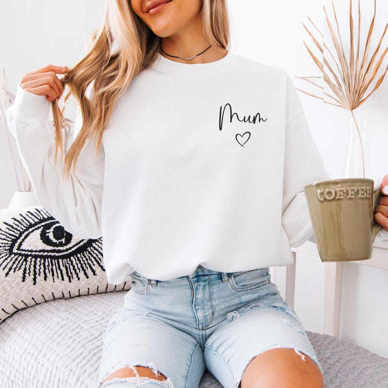 May include: A white sweatshirt with the word "Mum" and a heart symbol in black script. The person is holding a green mug with the word "Coffee" on it. The person is wearing light blue denim shorts.