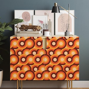 May include: A rectangular cabinet featuring a repeating pattern of interlocking circles in shades of orange, brown, and cream. The cabinet has two doors, a light wood top surface, and is supported by four thin, gold-coloured metal legs.