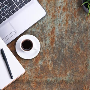 K&ouml;nnte beinhalten: Ein silberner Laptop, ein wei&szlig;es Notizbuch mit einem schwarzen Stift und eine wei&szlig;e Tasse mit schwarzem Kaffee. Die Gegenst&auml;nde sind auf einer texturierten Oberfl&auml;che mit verwitterter, rostiger Optik in Braun-, Orange- und Gr&uuml;nt&ouml;nen angeordnet.