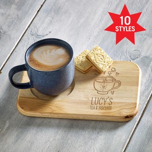 May include: A dark blue ceramic mug filled with coffee featuring latte art, placed on a rectangular wooden serving board with a circular indentation. The board is engraved with a tea cup graphic and the text "LUCY'S TEA & BISCUITS". Two square sandwich biscuits sit on the board.