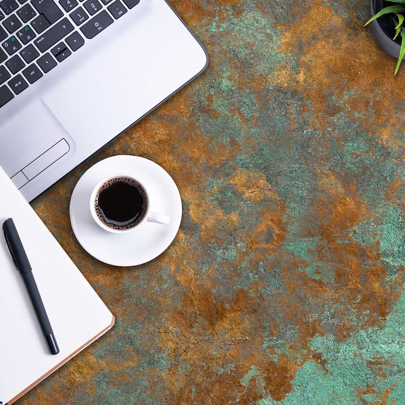 K&ouml;nnte beinhalten: Ein silberner Laptop, eine wei&szlig;e Kaffeetasse mit schwarzem Kaffee auf einer passenden Untertasse, ein wei&szlig;es Notizbuch und ein schwarzer Stift auf einer strukturierten Oberfl&auml;che mit einem gesprenkelten Muster aus oxidiertem Kupfergr&uuml;n und tiefem Metallic-Braun.