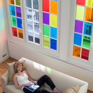 Pode incluir: Um quarto luminoso com grandes janelas com vitrais coloridos. As janelas s&atilde;o divididas em quadrados vermelhos, laranjas, amarelos, verdes, azuis e roxos. Uma mulher com uma blusa branca e cal&ccedil;as pretas relaxa em um sof&aacute; creme, segurando um livro.