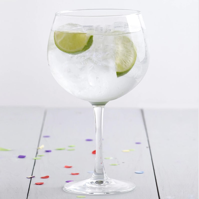 May include: A clear glass gin glass filled with ice and lime wedges. The glass is sitting on a white wooden surface with colorful confetti scattered around it.