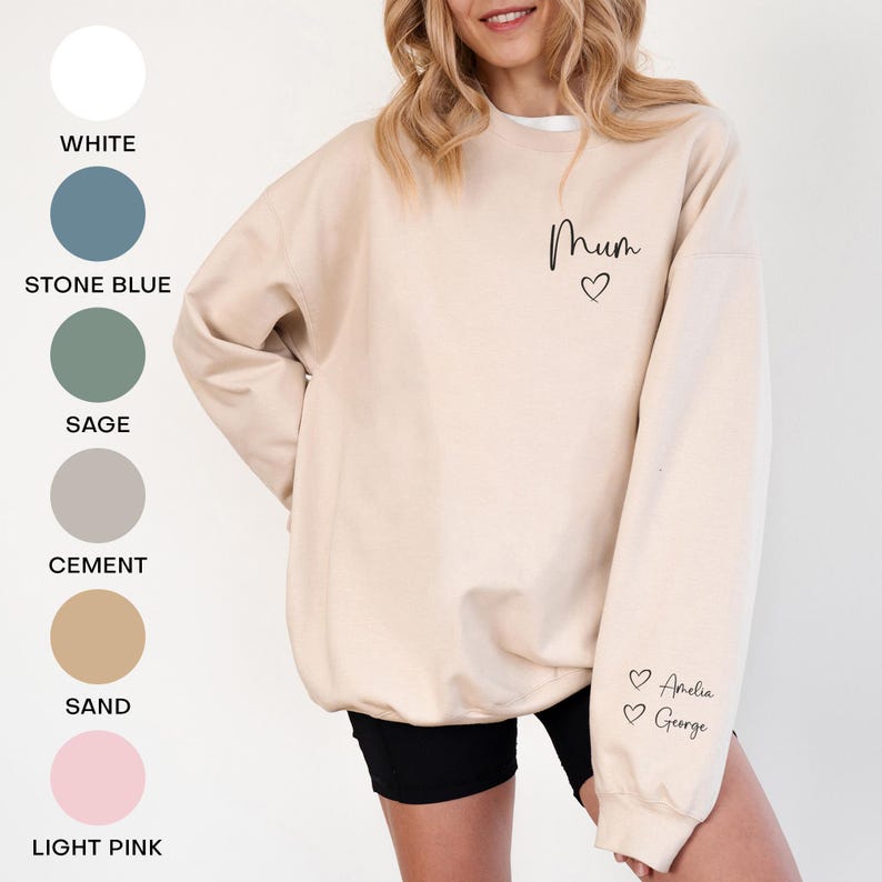 May include: A beige sweatshirt with the word "Mum" and a heart graphic on the chest. The sleeve features the names "Amelia" and "George" with heart symbols. Color swatches for white, stone blue, sage, cement, sand, and light pink are shown.