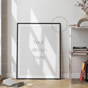 Könnte beinhalten: Ein schwarzer gerahmter Poster-Mockup mit dem Text "Your design here" auf weißem Hintergrund. Das Poster lehnt an einer weißen Wand mit einem Bücherregal dahinter. Das Bücherregal enthält Bücher und eine Vase mit getrockneten Blumen.