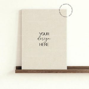 May include: A beige canvas print with the text "YOUR design HERE" in a stylish font. The print is displayed on a dark brown wooden shelf against a white wall. The image is a mockup.