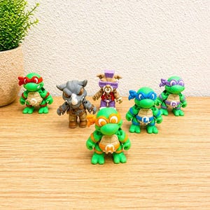 May include: A collection of colorful action figures of the Teenage Mutant Ninja Turtles and other characters. The figures are green, orange, blue, and purple, with various accessories and details. They are arranged on a wooden surface.