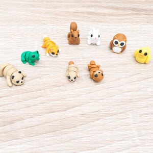 May include: A collection of small, colorful animal figurines. The assortment includes a panda, frog, fox, squirrel, rabbit, owl, chick, and two other unidentified animals. The figurines are made of a soft material and are arranged on a light wood-grain surface.