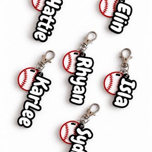 May include: Six personalized keychains featuring a white baseball with red stitching and a black acrylic name tag attached to a silver metal lobster clasp. The names Hattie, Elin, Karlee, Rhyan, Isla, and Sydney are displayed in white script.