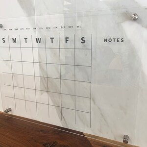 May include: A clear acrylic dry-erase calendar with a grid for each day of the month. The calendar has the months of the year listed across the top and the days of the week listed across the bottom. There is a section labelled "NOTES" on the right side of the calendar.