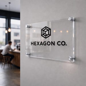 May include: A clear acrylic sign displaying the text "HEXAGON CO." in black. The sign has a geometric logo and is mounted on a grey wall with silver fixings. The sign is rectangular in shape and is likely for a business.