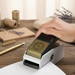 May include: A black and gold stamp, marked "MARK MAKER SAMPLE COMPANY", is being used on white paper. A hand presses the stamp. A gold pen and twine are visible in the background, on a wooden surface.