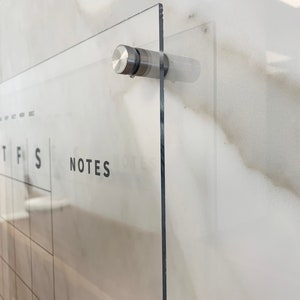 May include: A clear acrylic dry-erase calendar with a grid for each day of the week and a section for notes. The calendar is mounted to a wall with a silver metal screw.