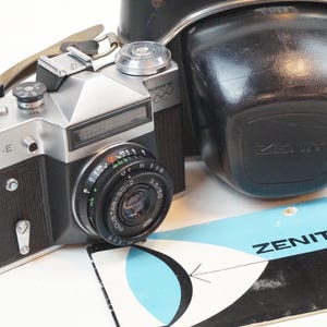 May include: A silver and black Zenit-E 35mm film camera with a black leather camera case. The camera has a black leather strap and a silver lens cap. The camera case has the word "Zenit" printed on it.